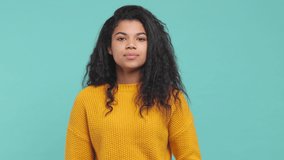 Smiling young african american woman 20s in yellow sweater isolated on blue turquoise background in studio. People lifestyle concept. Looking camera waving and greeting with hand as notices someone