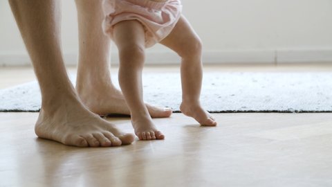 Barefoot infant learning walking with help of father. Side closeup view of Caucasian dad legs walking behind little baby and holding son or daughter. Making first step and family concept