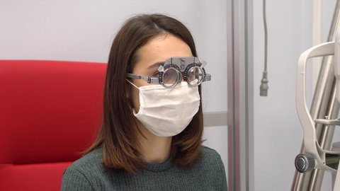 A young woman sits in special glasses for the selection of the necessary lenses at the eye doctor. The girl wears a protective mask from the virus.