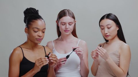Trio of beautiful diverse women applying nude makeup cosmetic products using lipstick and mascara. Beauty procedure. Makeup concept. Isolated.