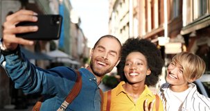 Close up portrait of mixed-races friends standing in city and making selfie photos on smartphone on sunny day. Joyful Caucasian guy taking pictures on street with women and African American female