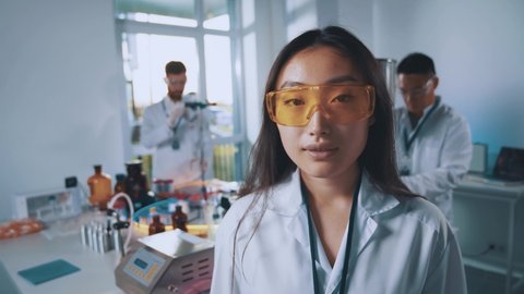 Portrait of young female scientist asian woman wearing white lab coat and protective eyeglasses conducting experiments with colleagues. Science and chemistry.