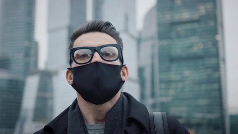 Man in eyeglasses and protective mask on background of business skyscrapers, looking up to the sky. Gimbal pan shot of businessman in coat standing near international business centre