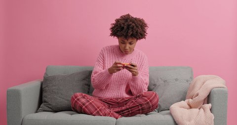 Female gamer holds modern mobile phone horizontally plays new game tries to pass level concentrated at smartphone display sits on comfortable sofa at home. Technology and online lifestyle concept