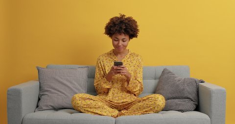Happy dreamy dark skinned woman sits in lotus pose on comfortable sofa uses smartphone for chatting or shopping online dressed in sleepwear being addicted to modern technologies reads social media