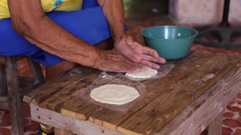 Hands of an elderly woman crushing a corn tortilla on a wooden bench, interior of an old house