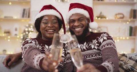 New normal and winter holidays. Close up shot of young black couple in Santa hats sitting at home with champagne and celebrating Christmas, congratulating friends via video call, web camera pov