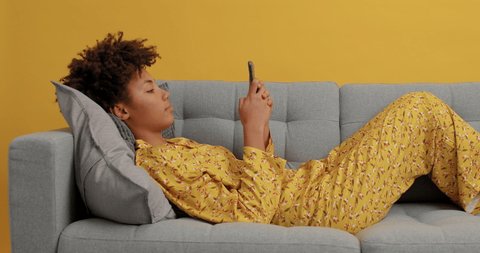 Lazy young Afro American woman uses mobile device enjoys chatting online wears yellow nightwear reads social media spends leisure time at home scrolls newsfeed sends text messages for friends