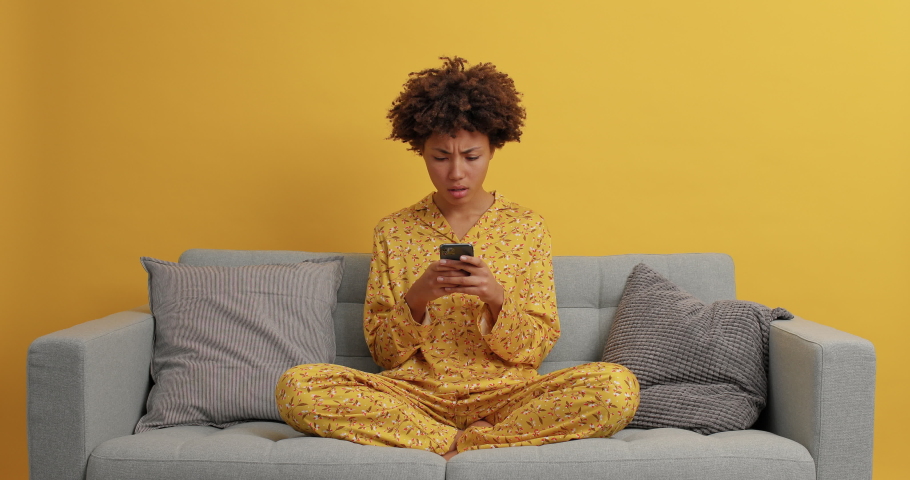 Displeased dark skinned woman looks angrily at smartphone types text messages sits crossed legs wears casual domestic clothes poses on sofa leaves comments under internet post has online communication Royalty-Free Stock Footage #1063133170