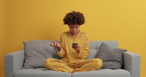 Displeased dark skinned woman looks angrily at smartphone types text messages sits crossed legs wears casual domestic clothes poses on sofa leaves comments under internet post has online communication