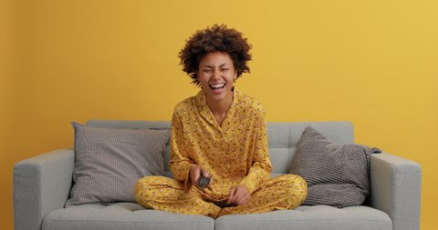 Young cheerful Afro American woman wears pajama spends time on quarantine poses on comfortable sofa switches channels on remote controller watches funny movie laughs happily enjoys lazy weekend