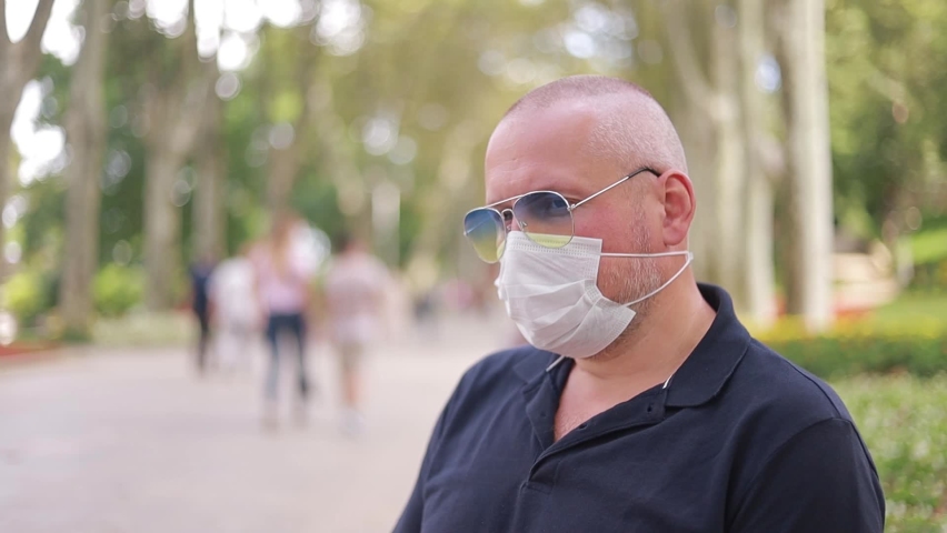 Emotional adult man in a protective white mask and blue sunglasses in a park outdoors on a summer day, defocus background.