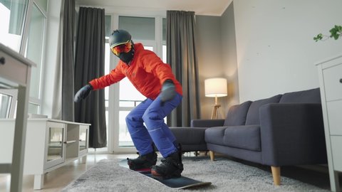 Fun video. Man dressed as a snowboarder rides a snowboard on a carpet in a cozy room. Waiting for a snowy winter