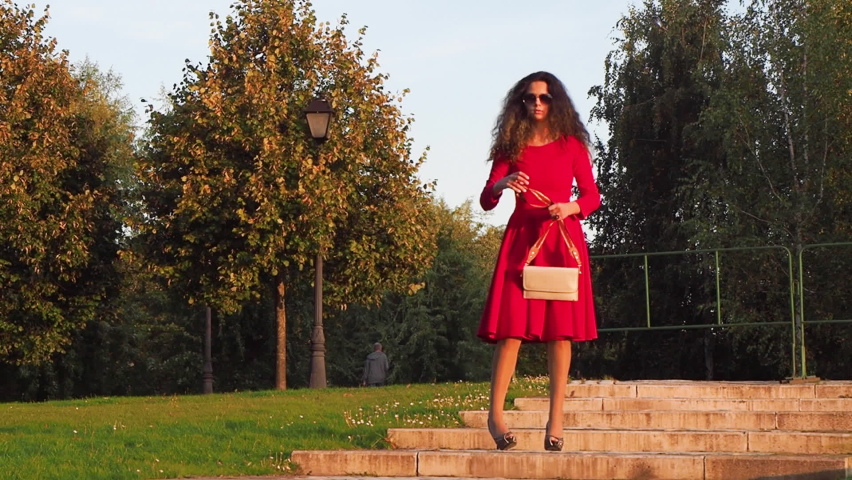 Drunk elegant lady in heels and a red dress is Walking through the city park after graduation and staggers from side to side