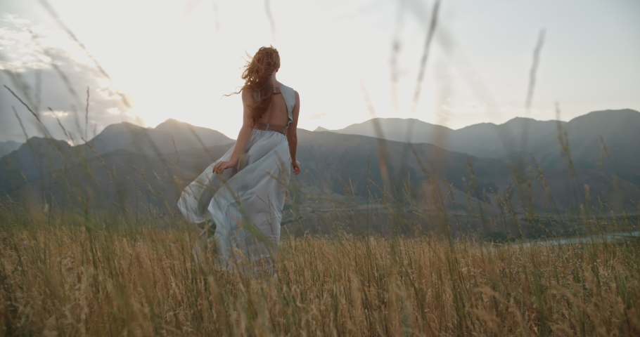 Young beautiful girl with red hair wearing white dress running on top of a mountain facing wind blowing her hair and dress and smiling - freedom, adventure, harmony 4k footage Royalty-Free Stock Footage #1063141084