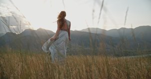 Young beautiful girl with red hair wearing white dress running on top of a mountain facing wind blowing her hair and dress and smiling - freedom, adventure, harmony 4k footage