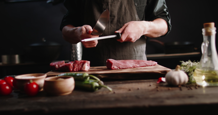 Chef sharpening his knife in front of kitchen table. Cooker preparing his tools before cutting raw pieceof meat and various vegetables 4k footage | Shutterstock HD Video #1063141135