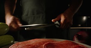 Chef sharpening his knife in front of kitchen table. Cooker preparing his tools before cutting raw piece of meat and various vegetables 4k footage