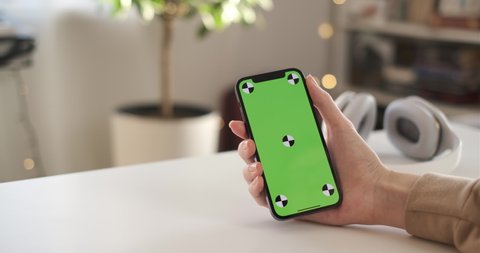 Russia - November 30 2020: Woman using smartphone iPhone 12 Pro vertical green screen. Female holding in hand portable gadget close up indoors living room. Mock-up for tracking or watching content
