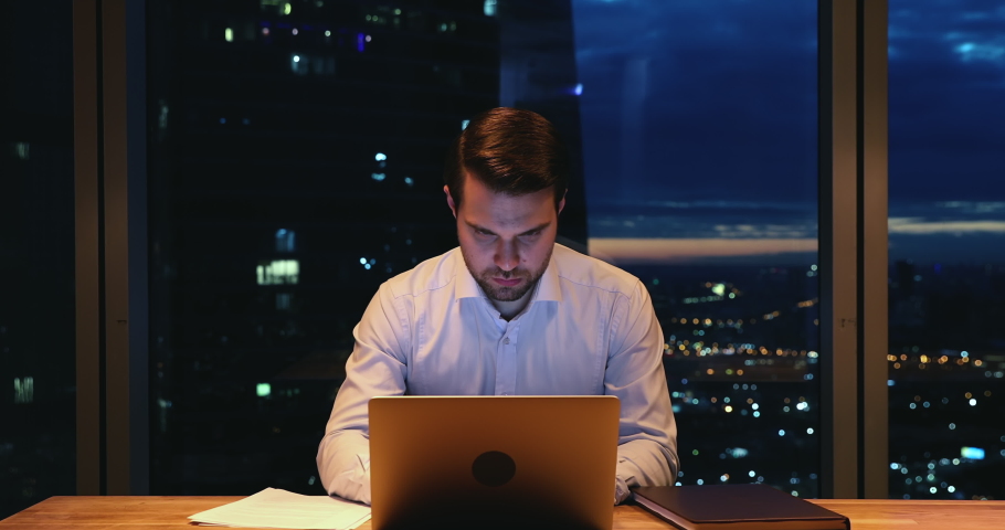 Serious focused 35s businessman work until late in office using laptop makes notes making urgent deadline task at modern workplace, night skyscrapers city view through window. Over hours work concept Royalty-Free Stock Footage #1063141570