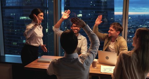 Team of young motivated ambitious startupper working until late in modern office boardroom, skyscrapers night city view through panoramic window. Accomplish successful meeting giving high five gesture