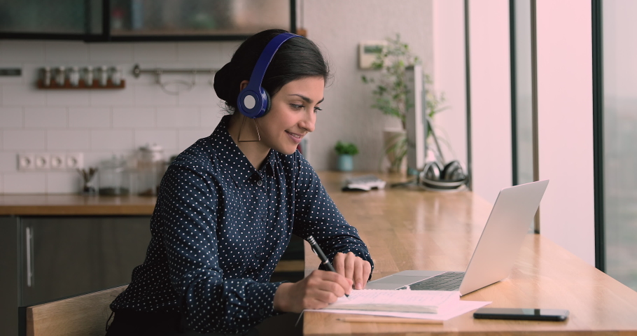 Indian millennial woman sit table in domestic kitchen wear headphones listen audio course english learn language using laptop and internet, writing notes memorizing information. Self-education concept | Shutterstock HD Video #1063141942