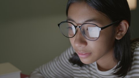 Asian little girl eyes in glasses looking at computer screen at home night. Close up of young face