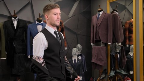 a man puts on a jacket in a clothing boutique in front of the mirror. Businessman choosing a suit in a luxury shop against the backdrop of suits, shoes and a mannequin.