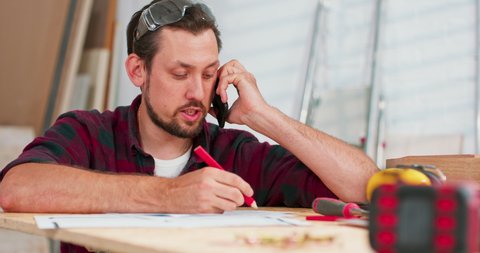 Portrait of modern young bearded man speaking by phone with client smiling and making notes in woodworking shop. The carpenter arranges the details of the order with the client, jokes and smiles.