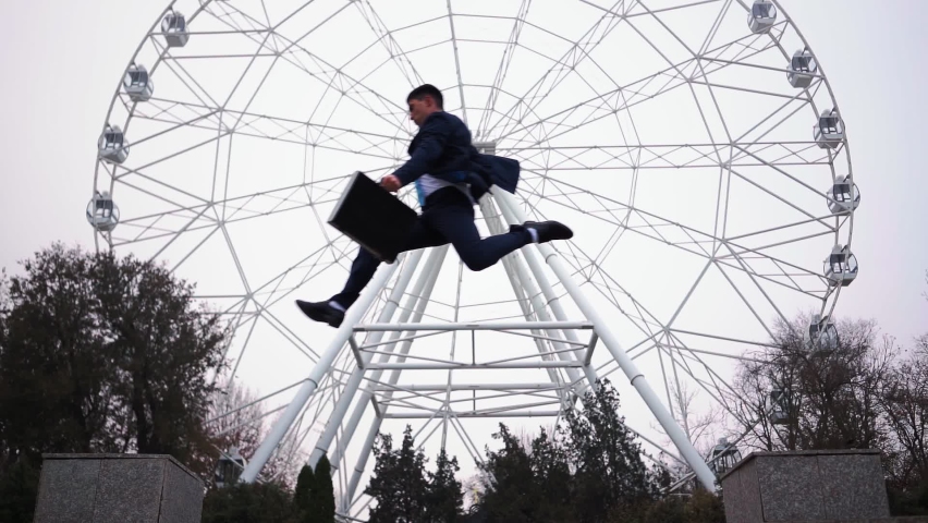 A successful man in a business suit and with a suitcase in his hands jumps over an obstacle against the background of a Ferris wheel in a city park. Parkour. Slow motion. High quality FullHD footage | Shutterstock HD Video #1063159795