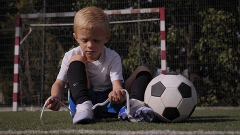 A little boy soccer player ties his shoelaces on a football field, he is sitting on the grass next to the ball.