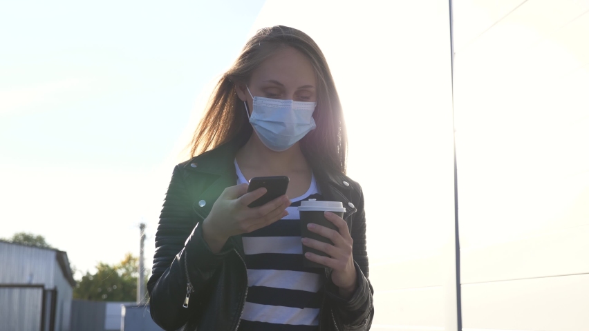 Girl student in a medical mask protection from infection in the park. A student uses a phone during a virus outbreak. A girl is standing in the park with a phone in a protective medical mask.