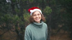 A young cheerful woman in a red Santa hat and a cozy warm sweater is recording a video with New Year and Christmas greetings against the background of pine trees in the forest.