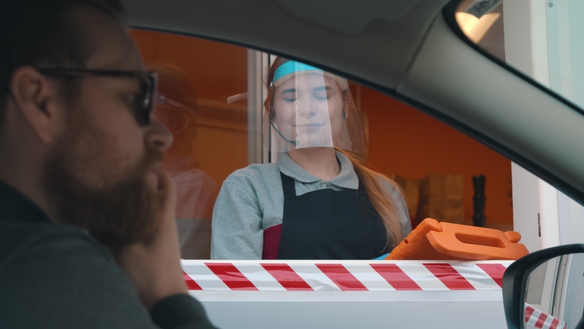 Bearded man driver in car paying with credit card for fast food order at drive thru counter. Young cafe employee in delivery window wearing protective shield and gloves holding terminal for payment Royalty-Free Stock Footage #1063163680