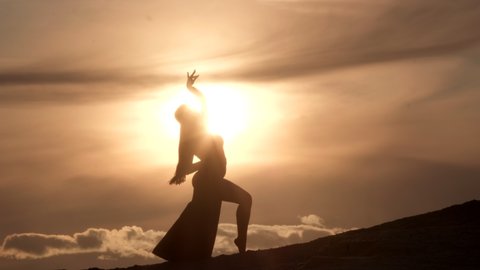 Silhouette of a woman dancing oriental belly dance on a sandy hill at sunset.