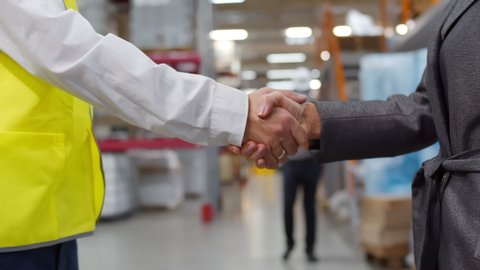 Closeup of businesswoman and industrial worker shaking hands on factory warehouse background. Industrial storehouse owner and manager handshaking after successful business meeting