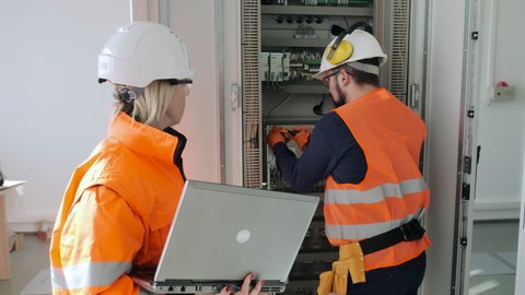 Skilled technician fixes switchboard in cabinet while female colleague controls process with laptop at electrical substation