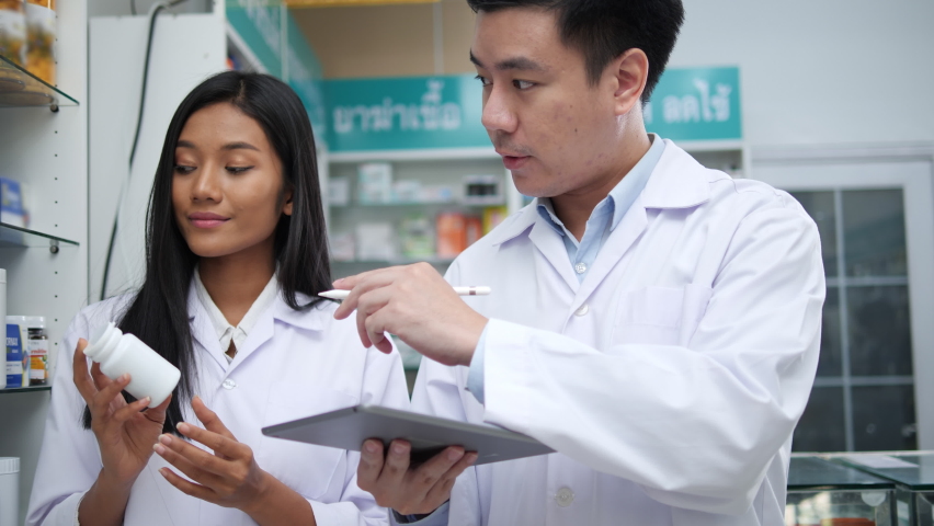 Asian pharmacists working and checking drug in pharmacy store at hospital. Asian doctor checking medicine cabinet and wearing white medical coat. Shelves pharmacy background in drugstore. | Shutterstock HD Video #1063168756