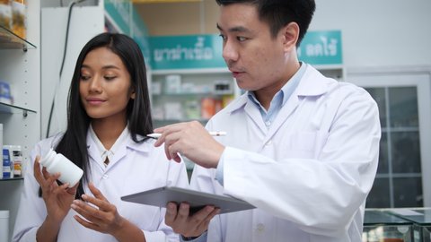 Asian pharmacists working and checking drug in pharmacy store at hospital. Asian doctor checking medicine cabinet and wearing white medical coat. Shelves pharmacy background in drugstore.