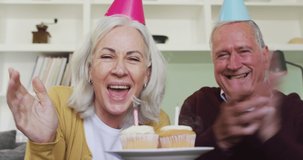 Senior couple celebrating a birthday making a video call, in party hats, the woman holding cupcakes, both blowing kisses and smiling, sitting at home. time together at home during a pandemic