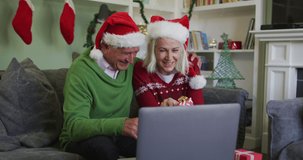 Happy senior caucasian couple celebrating christmas wearing santa hats, making a video call using a laptop, holding presents, sitting at home on sofa, in slow motion. quality time together at home dur