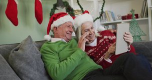 Happy senior caucasian couple celebrating christmas wearing santa hats, making a video call using a tablet computer, sitting at home on sofa, in slow motion. quality time together at home