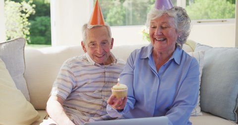Senior caucasian couple celebrating a birthday, wearing party hats, using laptop computer for video chat, woman holding cupcake with one lit birthday candle, sitting on sofa in sitting room 