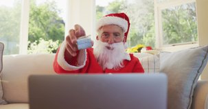 Senior caucasian man at christmas time  wearing santa costume, showing present, using laptop computer for video chat, sitting on sofa in living room in slow motion. enjoying christmas time at home