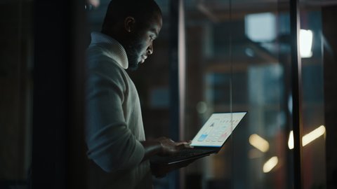 Handsome Black African American Male is Standing in Meeting Room Behind Glass Walls with Laptop Computer in an Creative Agency. Project Manager Wearing White Jumper and Working on App User Interface.