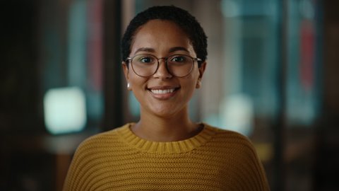 Close Up Portrait of a Young Latina with Short Dark Hair and Glasses Posing for Camera in Creative Office. Beautiful Diverse Multiethnic Hispanic Female Wearing Yellow Jumper is Happy and Smiling.