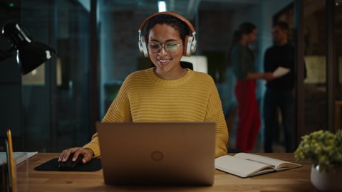 Young Hispanic Project Manager in Headphones and Working on Laptop Computer in Busy Creative Office Environment. Beautiful Diverse Multiethnic Female Specialist is Writing Business Strategy.