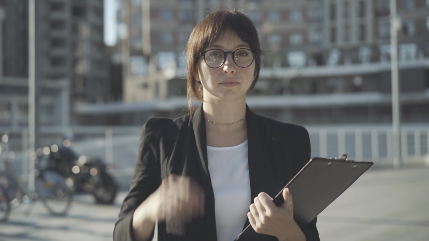Portrait of confident successful woman in eyeglasses standing on sunny city street and looking at camera. Elegant young Caucasian businesswoman posing outdoors. Confidence and success concept. | Shutterstock HD Video #1063174753