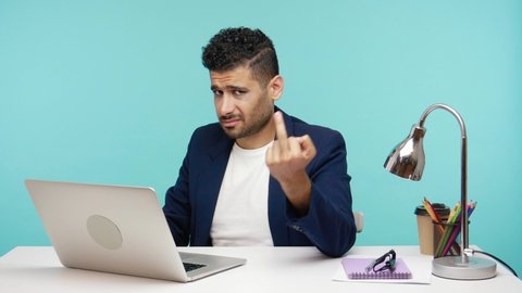 Self confident rude man showing middle finger fuck gesture at camera asking to get off, working on laptop. Indoor studio shot isolated on blue background
