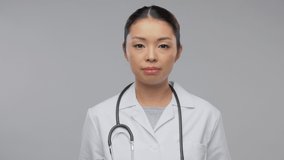 medicine, profession and healthcare concept - video portrait of happy smiling asian female doctor in white coat with stethoscope showing thumbs up over grey background